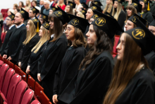 Grupa młodych ludzi. Stoją w rzędach, przed nimi i za nimi są czerwone fotele. Są ubrani w czarne togi i czarne birety z żółtym logo wydziału i chwostem. Są poważni, bo wygłaszają słowa przysięgi absolwenckiej.