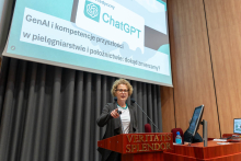 A middle-aged woman standing behind a lectern and giving a presentation. The woman is wearing a dark jacket and a white t-shirt. She wears glasses. Behind her you can see a projection screen with a blue slide. The woman has her right hand raised, gesturing.