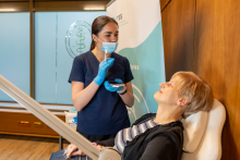 Dziewczyna w niebieskich rękawiczkach i granatowym uniformie stoi obok pacjentki  - to kobieta w średnim siedząca na fotelu dentystycznym. Dziewczyna trzyma narzędzie dentystyczne i małe lusterko. 