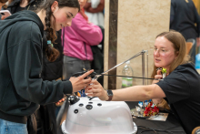 Dwie osoby biorące udział w interaktywnej demonstracji trenażera do zabiegów laparoskopowych. Osoba po lewej stronie jest ubrana w czarną bluzę z kapturem i dżinsy. Trzyma końcówki trenażera i ćwiczy na trenażerze. Osoba po prawej stronie jest ubrana w czarną koszulkę i nosi okular. Pomaga przy obsłudze trenażera.