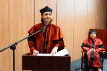 Kobieta w średnim wieku, prodziekan ds. analityki medycznej, stoi za mównicą i przemawia do mikrofonu na statywie. Czyta słowa przysięgi akademickiej. Jest ubrana w pomarańczowo-czarną togę, na głowie ma czarny biret. Za nią, w tle, widać jedną osobę siedzącą na fotelu, ubraną w togę akademicką.