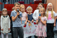 A group of children standing on the auditorium stage. Two children in the foreground are holding microphones and singing. Other children can be seen in the background holding a large, colorful poster with a drawing of a woman and red hearts. The poster reads “Thank you!” in red marker. The children are dressed in a variety of costumes.
