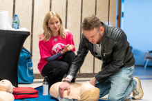 Dwie osoby ćwiczące resuscytację krążeniowo-oddechową (RKO) na manekinie. Osoba po prawej stronie, ubrana w czarną kurtkę i jasne dżinsy, klęczy na jednym kolanie i wykonuje uciski klatki piersiowej na manekinie. Osoba po lewej stronie, ubrana w różową koszulkę z białym nadrukiem, siedzi na piętach i obserwuje.