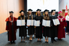 Grupa 7 osób. W środku absolwentki farmacji, młode kobiety, ubrane w togi. W rękach trzymają rozłożone teczki z dyplomami. Po lewej stoi dziekan, po prawej prorektorka. Wszyscy się uśmiechają. W tle, po prawej stronie widać flagę WUM. Za grupą widać też ekran projekcyjny.