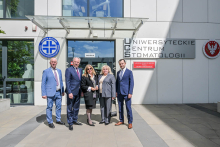 Pięć osób przed wejściem do budynku. Osoby są elegancko ubrane. Budynek ma białe ściany i duże szklane drzwi. Po prawej stronie drzwi widnieje napis: „Uniwersyteckie Centrum Stomatologii".