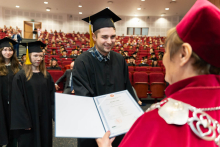 Młodzi ludzie, absolwenci, odbierający dyplomy od prorektorki. Na pierwszym planie jest młody mężczyzna, który uśmiecha się odbierając dyplom. Odbierany dyplom jest w rozłożonej teczce. Prorektorka trzyma go lewą dłonią.