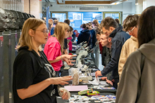 Grupa ludzi zgromadzonych wokół stołów z mikroskopami i innymi materiałami edukacyjnymi. Ludzie są zaangażowani w różne aktywności, niektórzy patrzą przez mikroskopy, inni rozmawiają lub przeglądają materiały na stołach.