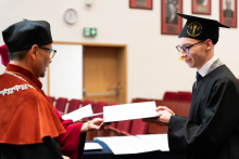 Młody absolwent, ubrany w togę i biret odbiera dyplom od dziekana. Na nosie ma okulary, pod szyją krawat. Mężczyźni są zwróceni do siebie przodem. W tle widać ścianę auli z powieszonymi na niej portretami.