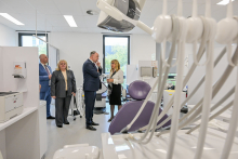 W pomieszczeniu przypominającym gabinet stomatologiczny są cztery osoby, które rozmawiają. Dwie osoby mają na sobie garnitury. Jedna osoba nosi ciemny strój. Czwarta osoba ubrana jest w jasną bluzkę i ciemną spódnicę. Na pierwszym planie widać fotele dentystyczne w kolorze fioletowym. Obok znajdują się białe narzędzia dentystyczne. W tle widoczne jest okno z widokiem na zewnątrz. W pomieszczeniu znajdują się też szafki i urządzenia medyczne.