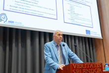 A man in a light blue jacket, shirt and striped tie. He stands behind a wooden lectern. Behind him is a large projection screen showing two slides of a presentation on dementia care and the JADE Health project co-funded by the European Union. 