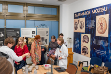 Grupa ludzi zgromadzonych wokół stołu na terenie muzeum. Jedna z osób ubrana jest w biały fartuch. W tle znajdują się duże plakaty z obrazami i tekstem na temat mózgu, zatytułowane "Tajemnice mózgu" Na stole znajduje się kilka słoików.