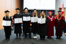 Grupa 7 osób. Od lewej stoją absolwenci farmacji, trzy młode kobiety i dwóch młodych mężczyzn, ubrani w togi. W rękach trzymają rozłożone teczki z dyplomami. Po prawej stoi prorektorka i dziekan. Wszyscy się uśmiechają. W tle, po prawej stronie widać flagę WUM. Za grupą widać też ekran projekcyjny.