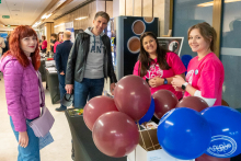 Grupa osób stojących przy stoisku. W centrum obrazu widoczny jest mężczyzna w skórzanej kurtce i szarej bluzie, który rozmawia z trzema kobietami. Dwie kobiety po prawej stronie mają na sobie różowe koszulki z nadrukiem, a kobieta po lewej stronie nosi fioletową kurtkę i trzyma torebkę. Na pierwszym planie znajdują się balony w kolorach bordowym i niebieskim.