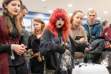 Grupa osób obserwuje młodą kobietę o intensywnie czerwonych włosach i wyrazistym makijażu, która obsługuje trenażer do zabiegów laparoskopowych. Osoba ta ma na sobie czarną bluzę z grafiką i skupia się na urządzeniu, trzymając uchwyty przypięte do przewodów. W tle widać zainteresowane twarze innych uczestników wydarzenia.