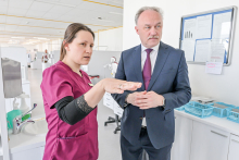 Dwie osoby stojące w jasnym pomieszczeniu. Pomieszczenie przypomina gabinet stomatologiczny. Osoba po lewej stronie nosi bordowy strój medyczny i czarną koszulkę. Osoba po prawej stronie ma na sobie ciemny garnitur, białą koszulę i bordowy krawat. W tle widać urządzenia medyczne.