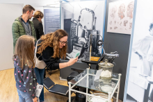 Grupa czterech osób oglądających wystawę muzealną. Na pierwszym planie znajduje się kobieta z długimi, falującymi włosami, która trzyma telefon i robi zdjęcie eksponatu. Obok niej stoi dziewczynka w szarej bluzce w kolorowe kropki oraz mężczyzna w zielonej kurtce. W tle widoczny jest jeszcze jeden mężczyzna. Eksponat to stary sprzęt medyczny umieszczony na szklanym stoliku z metalową ramą, na którym znajdują się różne przyrządy medyczne oraz pojemnik z watą. 