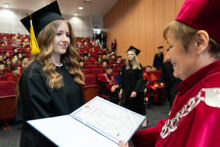 Młoda kobieta, absolwentka, ubrana w togę i biret odbiera dyplom od prorektorki. Absolwentka ma długie kręcone włosy. Uśmiecha się do prorektorki. Jest do niej zwrócona przodem. Prorektorka również się uśmiecha. W tle widać innych absolwentów, którzy idą odebrać dyplom. 