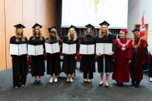 Grupa 8 osób. Od lewej stoją absolwentki farmacji, młode kobiety, ubrane w togi. W rękach trzymają rozłożone teczki z dyplomami. Po prawej stoi prorektorka i dziekan. Wszyscy się uśmiechają. W tle, po prawej stronie widać flagę WUM. Za grupą widać też ekran projekcyjny.