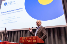 Mężczyzna w garniturze, stojący za mównicą z napisem "Veritas Splendor". Mężczyzna trzyma mikrofon i gestykuluje ręką. W tle widoczny jest ekran z prezentacją, na której znajduje się logo Uniwersytetu Mikołaja Kopernika w Toruniu.