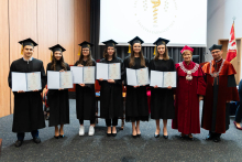 Grupa 8 osób. Od lewej stoją absolwenci farmacji, jeden młody mężczyzna i 5 młodych kobiety, ubranych w togi. W rękach trzymają rozłożone teczki z dyplomami. Po prawej stoi prorektorka i dziekan. Wszyscy się uśmiechają. W tle, po prawej stronie widać flagę WUM. Za grupą widać też ekran projekcyjny.