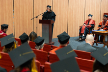 Kobieta ubrana w czarną togę i biret stoi na środku auli za mównicą i przemawia do mikrofonu. Na pierwszym planie zdjęcia widać absolwentów, którzy patrzą na mówczynię. Widać ich odwrócone głowy w biretach z chwostami.