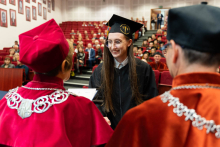 Młoda kobieta, absolwentka, ubrana w togę i biret odbiera dyplom od prorektorki i dziekana. Absolwentka ma długie ciemne włosy. Uśmiecha się. Prorektorka i dziekan są ujęci od tyłu, widać ich plecy, na szyjach mają łańcuchy akademickie oraz birety na głowach. W tle widać czerwone fotele i siedzące na nich osoby.