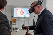 Dwaj mężczyźni w garniturach. Jeden z nich ma na głowie czarne gogle VR i trzyma czarne kontrolery VR. Drugi mężczyzna patrzy na ekran. Na ekranie widać wnętrze ust – zęby i język. W tle znajduje się krzesło i białe ściany.