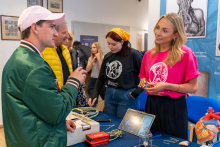 Na pierwszym planie po lewej stronie widać mężczyznę w zielonej kurtce i białej czapce, który rozmawia z kobietą po prawej stronie, ubraną w różową koszulkę z grafiką anatomiczną serca. Obok niej stoi inna kobieta w czarnej koszulce z podobną grafiką oraz żółtą chustką na głowie. Widać też stół, na którym znajdują się różne przedmioty, takie jak modele anatomiczne serca, laptop, kable i inne urządzenia elektroniczne. 