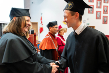 Po lewej kobieta w średnim wieku ubrana w czarną togę i biret. Po prawej młody mężczyzna w czarnej todze. Trzymają się za dłonie w geście gratulacji. Uśmiechają się, patrząc wprost na siebie. W tle widać dziekana.