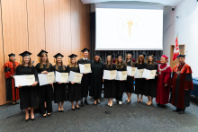 Grupa kilkunastu osób. Od lewej stoją absolwenci farmacji ubrani w togi. W rękach trzymają rozłożone teczki z dyplomami. Po prawej stoi prorektorka i dziekan. Wszyscy się uśmiechają. W tle, po prawej stronie widać flagę WUM, a po lewej prodziekan w todze. Za grupą widać też ekran projekcyjny.