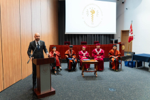 Grupa osób na środku auli. Po lewej mężczyzna w średnim wieku, Główny Inspektor Farmaceutyczny, ubrany w szary garnitur i krawat, stoi za mównicą i przemawia. Za nim na krzesłach siedzą władze dziekańskie i rektorskie ubrane w togi. W tle widać ekran projekcyjny i szare zasłony. Po prawej stoi flaga WUM.