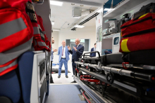 W tle, na środku zdjęcia, znajdują się trzy osoby w garniturach, które rozmawiają. Twarze tych osób są zamazane. Po prawej stronie widoczne są nosze z czarnym materacem. Na noszach leży czerwona torba ratunkowa. Wnętrze karetki jest dobrze oświetlone białym światłem sufitowym.