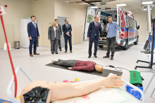 Pięć osób stojących w pomieszczeniu. W tle widać otwartą karetkę pogotowia. Wnętrze karetki jest dobrze widoczne. Na pierwszym planie leży manekin na czarnej macie. Manekin ubrany jest w bordową bluzę i ciemne spodnie. Obok manekina znajduje się sprzęt medyczny, w tym defibrylator. Po lewej stronie, na białym stole, leży sztuczna ręka z bandażem i śladami czerwonego barwnika.