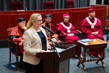 Na pierwszym planie za mównicą stoi kobieta w średnim wieku i przemawia. Ma okulary, jasne włosy, nosi jasną marynarkę i ciemną sukienkę. Za nią na krzesłach siedzą władze dziekańskie i rektorskie.