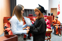 Po lewej kobieta w niebieskiej bluzce, z długimi włosami i okularach, po prawej absolwentka w czarnej todze z rudymi włosami. Kobieta po lewej wręcza absolwentce dyplom. Ściskają sobie dłonie w geście gratulacji. W tle widać inne osoby, władze rektorskie i dziekańskie.