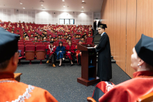 Młody mężczyzna ubrany w togę akademicką - przedstawiciel absolwentów, stoi za mównicą i przemawia. Jest zwrócony twarzą do zdjęcia, patrzy na władze rektorskie i dziekańskie. Za nim, na czerwonych fotelach siedzą uczestnicy uroczystości.