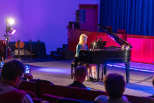 Kobieta gra na fortepianie koncertowym na oświetlonej scenie w ciemnej sali audytoryjnej. Artystka jest podświetlona fioletowym i niebieskim światłem scenicznym, a w pierwszym planie widoczna jest publiczność siedząca w czerwonych fotelach, z kilkoma osobami nagrywającymi występ telefonem.