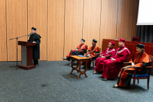 Młody mężczyzna, przedstawiciel samorządu, w czarnej todze stoi za brązową mównicą i przemawia. Za nim, na krzesłach siedzą władze dziekańskie i rektorskie.