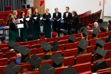 Grupa osób w togach i biretach siedzi na czerwonych krzesłach w audytorium. Na scenie w tle stoją osoby przemawiające lub śpiewające. Widoczny jest stół z niebieskim obrusem i flagi.