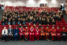 Duża grupa osób w togach i biretach siedzi w audytorium. W pierwszym rzędzie osoby w togach o różnych kolorach, za nimi absolwenci z dyplomami. Audytorium pełne, na schodach po bokach stoją ludzie.