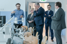 Pięć osób ubranych formalnie, które stoją w jasnym pomieszczeniu z symulatorami medycznymi. Ściany są pomalowane na biało i niebiesko. Na pierwszym planie leżą manekiny medyczne do ćwiczeń dentystycznych. Manekiny znajdują się na stołach, a nad nimi wiszą lampy. W tle widać drzwi.