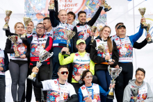 Grupa osób stojących na podium. Osoby trzymają puchary. Wszyscy mają na sobie kolorowe koszulki z nadrukami i numerami sportowymi. Na szyjach widoczne są medale. Tło jest jasne, z napisami i grafiką.
