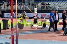 A female athlete wearing a yellow T-shirt with the number 1070 and gray shorts performs a javelin throw in the athletics stadium. In the background you can see people, sports equipment, stands and banners with the words “Ministry of Sports and Tourism” and “Academic Championships of Poland.”