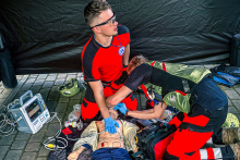 Dwóch ratowników medycznych w czerwono-czarnych uniformach wykonuje resuscytację na manekinie. Jeden prowadzi uciski klatki piersiowej, drugi używa worka samorozprężalnego. Obok leży defibrylator i sprzęt medyczny. Scena odbywa się na zewnątrz, pod czarnym namiotem.