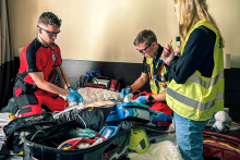 Dwóch ratowników medycznych w czerwonych uniformach udziela pomocy manekinowi leżącemu na łóżku. Obok stoi kobieta w żółtej kamizelce z napisem „Służba”, trzymająca długopis i notes. Wokół łóżka widoczne są torby ratownicze, defibrylator i kaski.