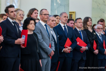 Grupa osób w eleganckich strojach stoi w rzędzie. Każda osoba trzyma czerwoną teczkę z godłem Polski. Tło jest jasne, z marmurowymi kolumnami i obrazem na ścianie.