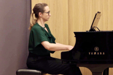 Kobieta ubrana w czarne spodnie i zieloną bluzkę siedzi przy fortepianie i gra. Fortepian jest czarny.