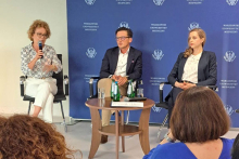 Trzech prelegentów siedzi na scenie przed niebieskim tłem z logo Warszawskiego Uniwersytetu Medycznego. Jeden z nich trzyma mikrofon. Przed nimi stolik z wodą. W tle widoczni uczestnicy siedzący tyłem do aparatu.