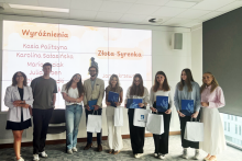 Osiem elegancko ubranych osób stoi w rzędzie, trzymając białe torby i niebieskie teczki. W tle ekran z napisem „Wyróżnienia” i „Złota Syrenka” oraz listą nazwisk. Pomieszczenie jasne, z dużym oknem po prawej stronie.