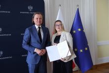 Dwie osoby w eleganckich strojach stoją obok siebie. Osoba po lewej ma niebieski garnitur, białą koszulę i krawat. Osoba po prawej trzyma otwartą teczkę z dokumentem. W tle granatowa ścianka z napisem „WOJEWODA MAZOWIECKI”, godłem Polski oraz flagi Polski i Unii Europejskiej.